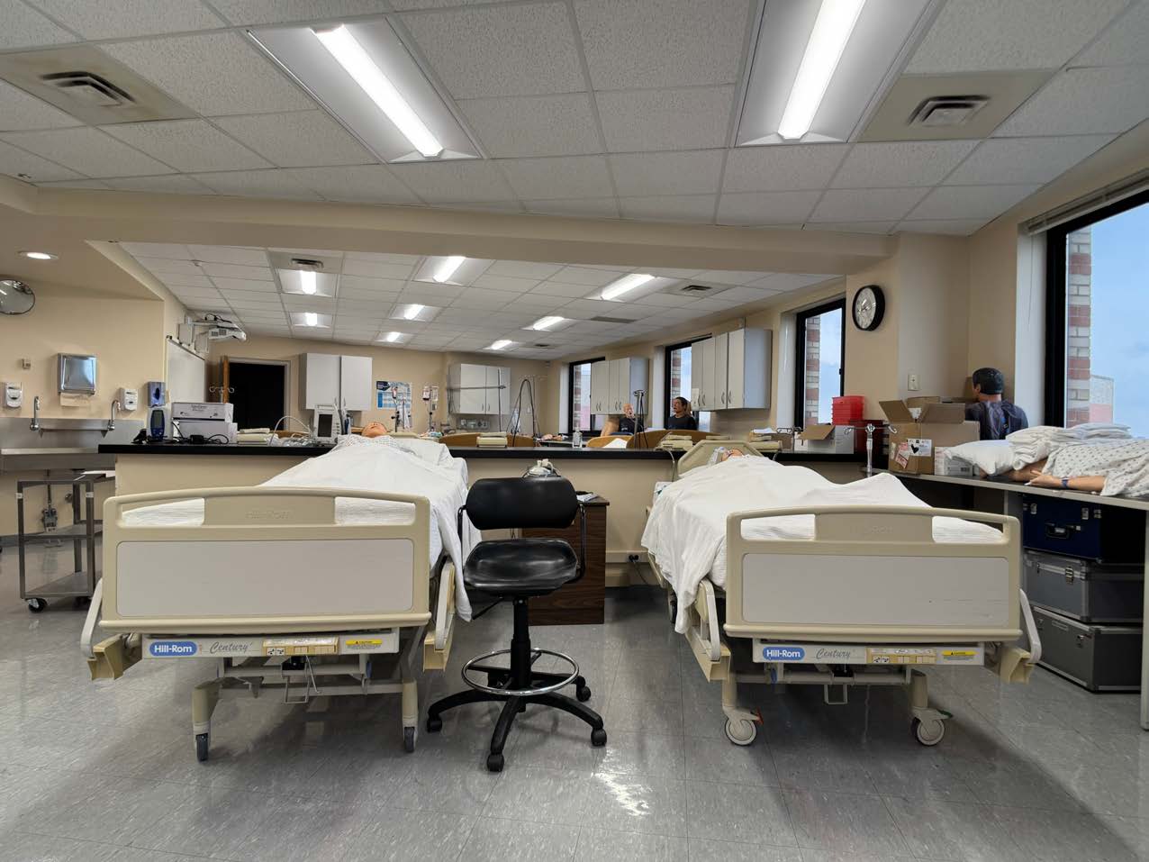 Photo of a nursing department classroom with two hospital beds containing medical dummies and a couple of students in the background.