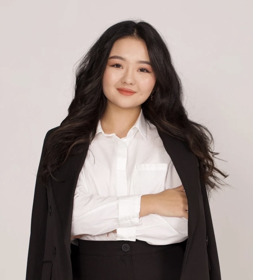 Smiling young woman with long, wavy, dark hair, a black coat over her shoulders, wearing a white blouse and black bottoms with her arms crossed
