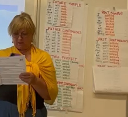 Person in front of whiteboard A person wearing a yellow shawl reading from a peice of paper in front of a classroom with class materials displayed on the whiteboard and wall in the background