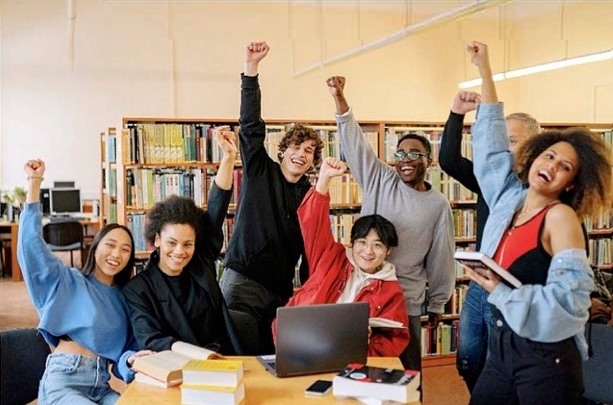 Students around a table with books A group of diverse students in a room with book shelves, sitting and standing around a table with a laptop and books on it, smiling and throwing their fists in the air
