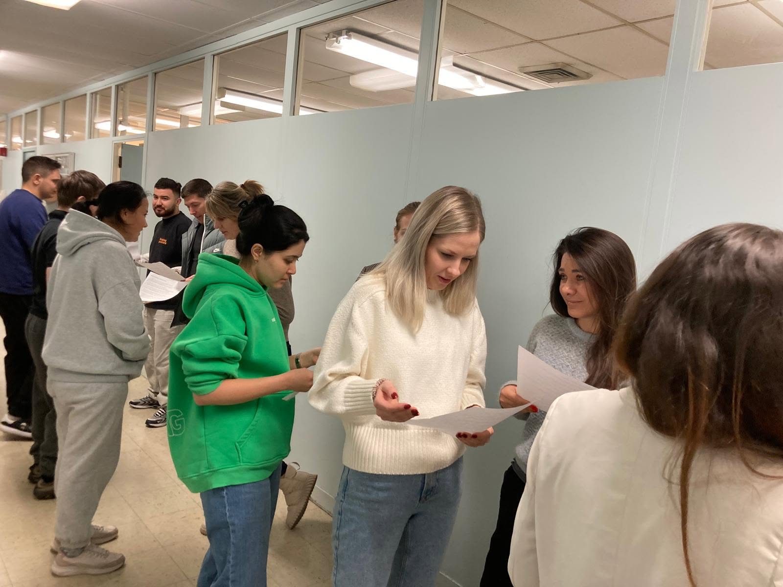 Students engaged in a conversation activity Students standin in pairs along a wall holding papers engaged in a conversation activity