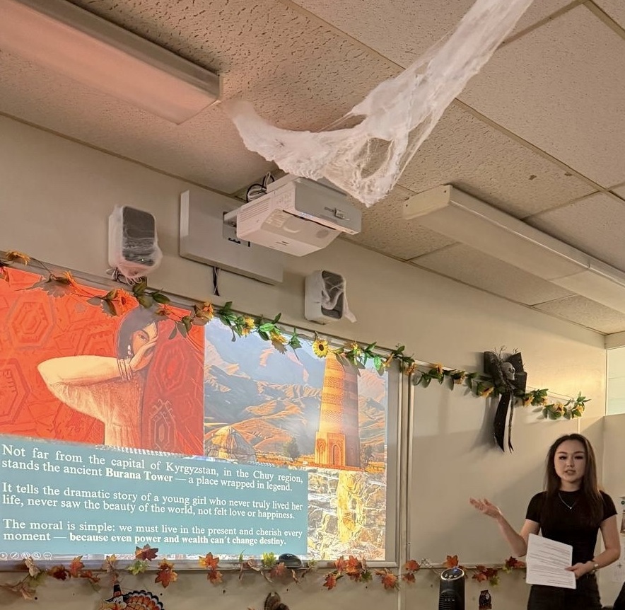 Young woman with long, dark hair wearing a black t-shirt front of a whiteboard, onto which a presentation about Kyrgyzstan is projected