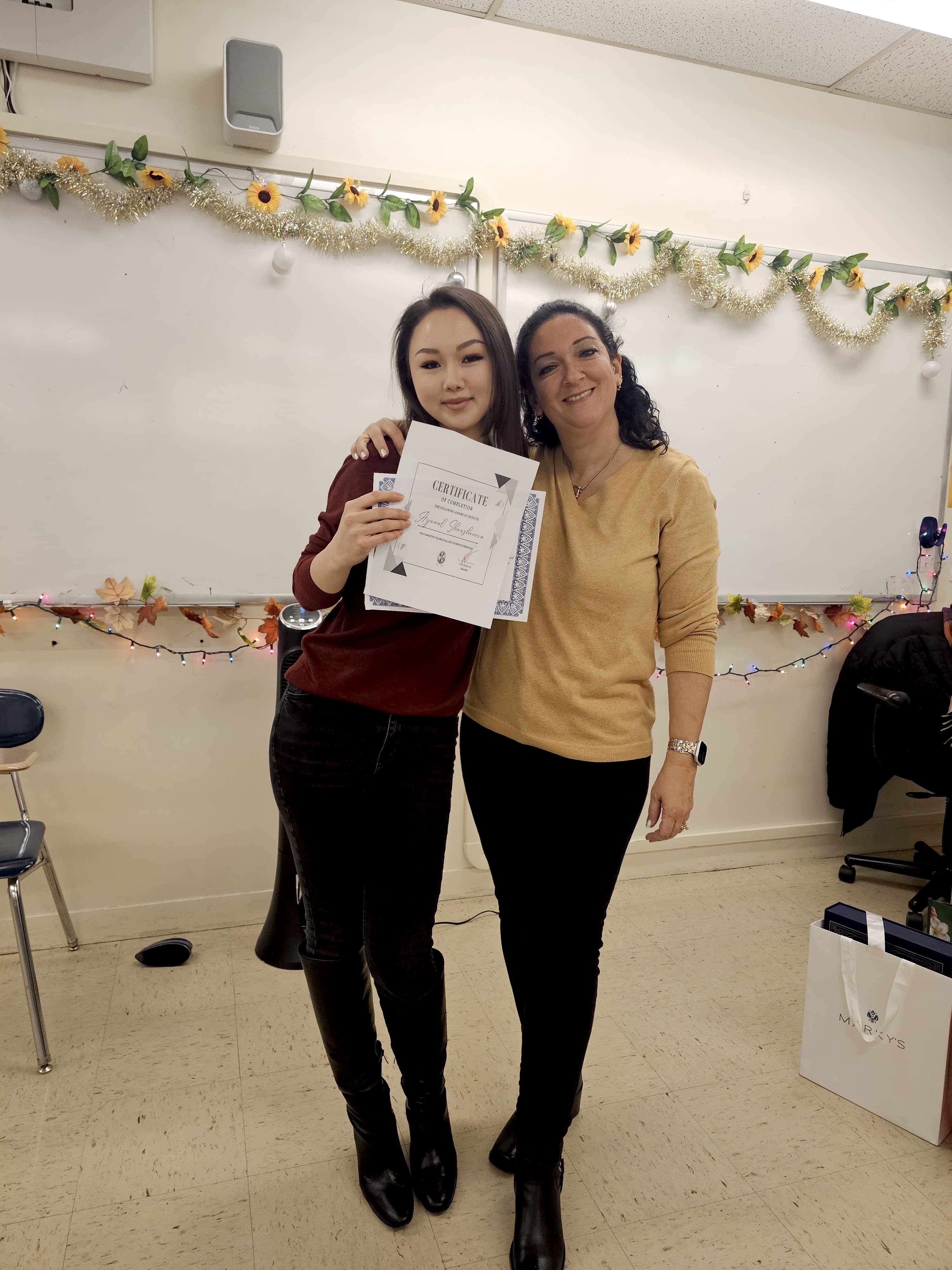 Two women with shoulder-length dark hair hugging in front of a whiteboard, one is holding up a diploma and a certificate