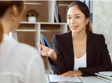 A smiling young woman with long black hair tied back, wearing a white v-neck top and a black blazer, sitting at a table with a clipboard in front of her, speaking to the person sitting across.