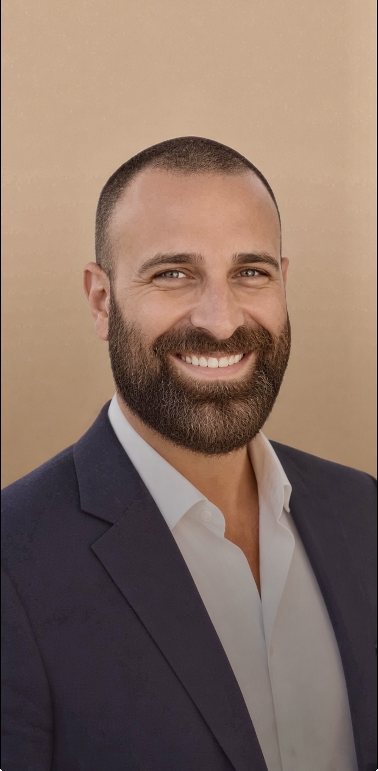 Smiling young man with a short, light brown beard in a dark blue blazer and white button down shirt