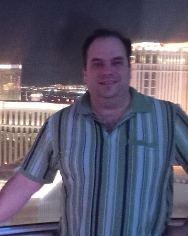 Smiling man in a striped polo shirt with tall, brightly lit buildings against dark sky behind him.