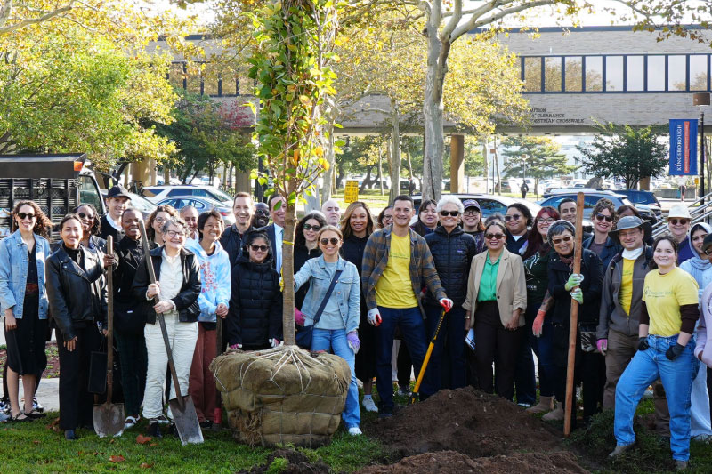 A large group of KCC community members, including President Duitch, stand with shovels around a freshly dug hole in the ground and a ready-to-plant tree outside the A building.
